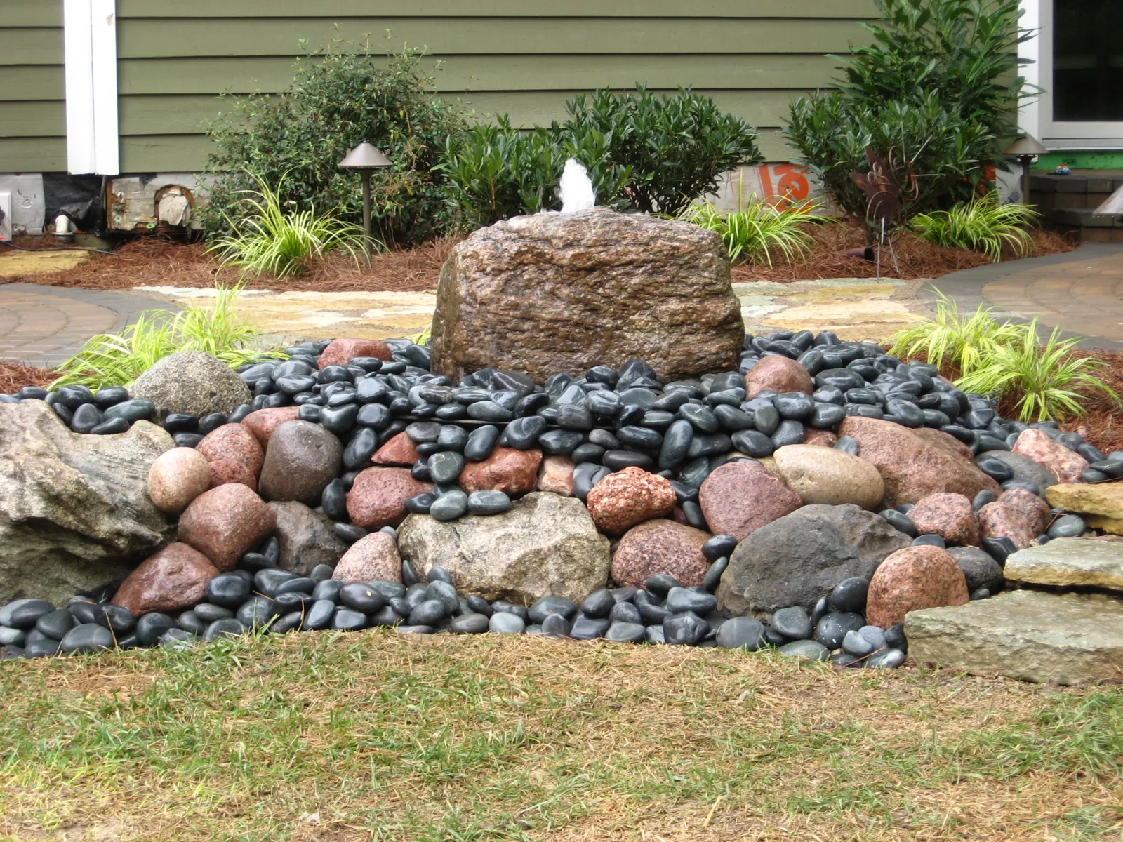 Drilled granite boulder fountain on black river rock with grasses